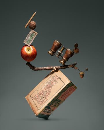 Absurd picture where a series of objects, including an apple, a pencil, a book, a pair of binoculars etc are impossibly balanced on top each other to prompt the viewer's interest.